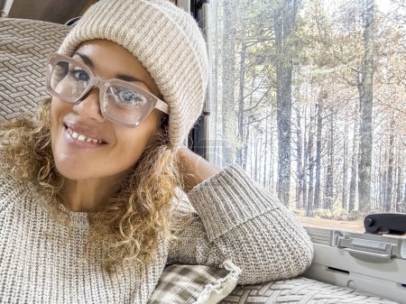 Foto de Hermosa mujer con sombrero de lana mira a la cámara sonriendo. Señora sentada en la caravana disfrutando de sus vacaciones en el bosque - Imagen libre de derechos
