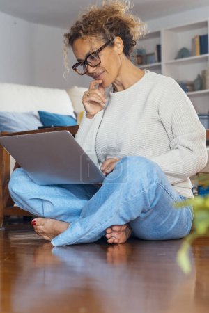 Téléchargez les photos : Femme attrayante travaillant sur ordinateur portable à la maison assis pieds nus sur le sol en pensant et en regardant l'écran. Les gens modernes et l'activité de loisirs en ligne dans le salon. Style de vie positif - en image libre de droit
