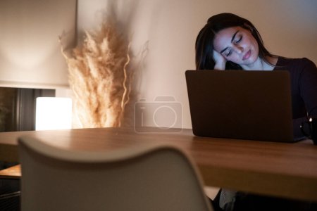 Young pretty smiling woman using laptop in home at night. Happy girl looking at computer. Cheerful people sitting at desk in living room. Concept of enterprising and hardworking person.