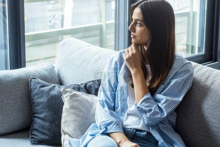 Photo for Thoughtful young woman with sad expression sitting on sofa at home looking outside and thinking bad problems on life. Lonely depressed adult female. - Royalty Free Image