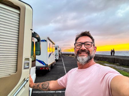  hombre feliz tomando foto selfie en el lado de una caravana moderna autocaravana. Viajero vanlife estilo de vida personas que comparten en las redes sociales aventura y viaje. Increíble puesta de sol en el fondo. Aparcamiento