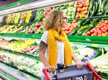 Foto de Hermosa mujer sonriente empujando carrito de la compra y frutas de los estantes en el supermercado. Comprar comida en la tienda de comestibles. - Imagen libre de derechos