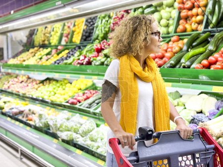 Foto de Hermosa mujer sonriente empujando carrito de la compra y frutas de los estantes en el supermercado. Comprar comida en la tienda de comestibles. - Imagen libre de derechos