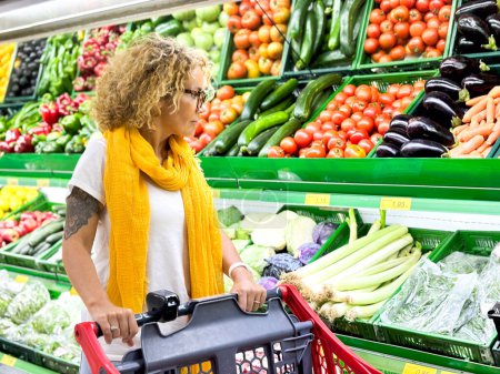 Photo for Beautiful smiling woman pushing shopping cart and  fruits off the shelves in supermarket. Buying food at grocery store. - Royalty Free Image
