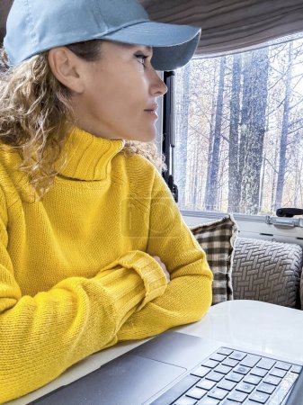 Téléchargez les photos : Belle femme au chapeau bleu et pull jaune regarde par la fenêtre du camping-car devant l'ordinateur portable. Dame aime la forêt assis de son camping-car - en image libre de droit