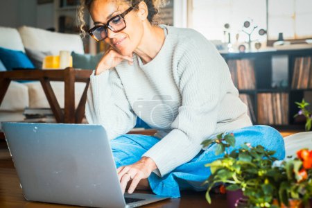 Téléchargez les photos : Femme adulte travaillant sur ordinateur portable seul à la maison assis sur le sol en bois. Connexion et technologie sans fil. Femmes modernes utilisant l'ordinateur pour rechercher sur le web et faire du shopping sur Internet en ligne - en image libre de droit