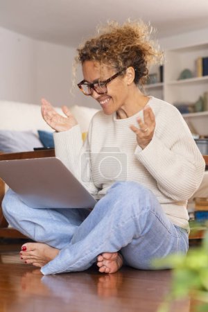 Téléchargez les photos : Excité femme heureuse faisant appel vidéo en utilisant un ordinateur portable à la maison assis pieds nus sur le sol. geste féminin avec les mains devant un ordinateur et une vidéoconférence. Réunion en ligne sortir ensemble site - en image libre de droit