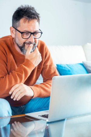 Photo for Adult freelance man looking the laptop screen thinking and touching chin. Wearing glasses mature male. Homework smart working remote job business concept lifestyle. Modern worker sitting on sofa - Royalty Free Image