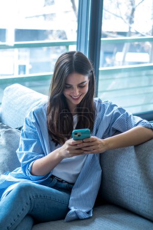 Happy girl scrolling and checking social media while holding smartphone at home. Smiling young brunette woman using mobile phone app to play games, shop online, order delivery while relaxing on sofa.