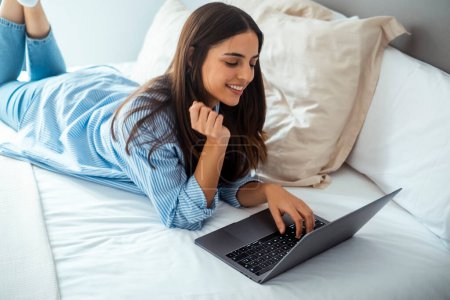  smiling woman lying down the bed in front of her laptop 