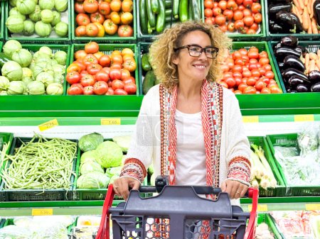 Foto de Hermosa mujer sonriente empujando el carrito de la compra y tomando frutas de los estantes en el supermercado. Comprar comida en la tienda de comestibles. - Imagen libre de derechos