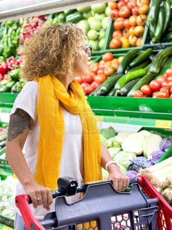 Téléchargez les photos : Belle femme souriante poussant chariot d'achat et de prendre des fruits sur les étagères dans le supermarché. Acheter de la nourriture à l'épicerie. - en image libre de droit