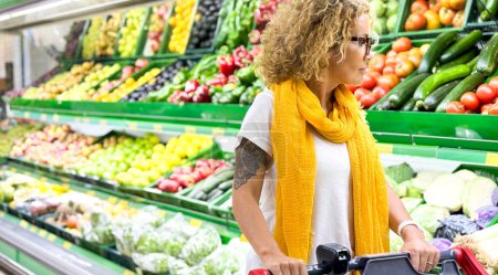 Téléchargez les photos : Belle femme souriante poussant chariot d'achat et de prendre des fruits sur les étagères dans le supermarché. Acheter de la nourriture à l'épicerie. - en image libre de droit