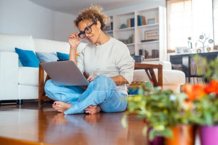 Photo for Middle age lady sitting on the floor comfortable using laptop at home in indoor technology wireless leisure activity alone. Modern female people working on computer. Woman browsing web and writing - Royalty Free Image