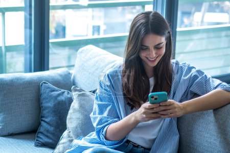 Happy girl scrolling and checking social media while holding smartphone at home. Smiling young brunette woman using mobile phone app to play games, shop online, order delivery while relaxing on sofa.