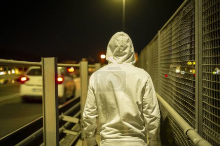 Photo for Back view of person with hoodle walking by night on the road with traffic cars on the street. Urban lifestyle people concept. Security and crime. Walk on a bridge - Royalty Free Image