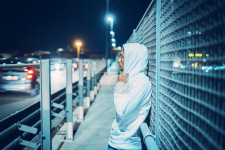 Side view of person with hoodie standing by night on the road with traffic cars on the street. Urban lifestyle people concept. Security and crime. Thoughts and loneliness