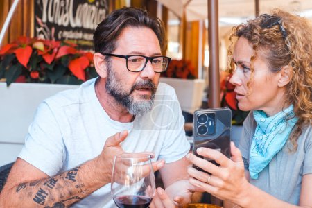Photo for Modern adult couple look on phone contents sitting at the restaurant table with worried expression. Woman show cell to a man. Unhappy tourist writing a bad review after lunch. Problems notification - Royalty Free Image