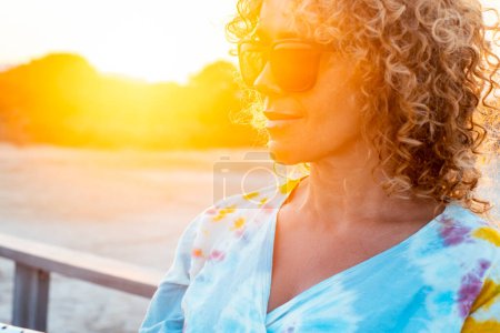 Photo for Sunny outdoor portrait of curly woman smiling and wearing sunglasses with sunset backlight. Golden portrait of attractive lady in outdoors leisure activity alone. Tourist happy. Beautiful female - Royalty Free Image
