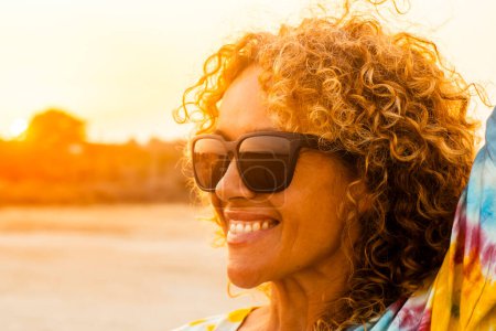 Photo for Sunny outdoor portrait of curly  woman smiling and wearing sunglasses with sunset backlight. Golden portrait of attractive lady in outdoors leisure activity alone. Tourist happy. Beautiful female - Royalty Free Image