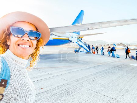 Téléchargez les photos : Une femme de 50 ans portant un chapeau et des lunettes de soleil prend du selfie avant de monter dans l'avion. Heureuse femme seule prend une photo souvenir. - en image libre de droit