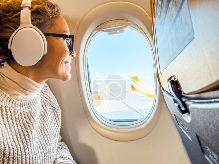 Lächelnder Passagier mit Bluetooth-Kopfhörer, der über die WLAN-Internetverbindung an Bord aus dem Bullauge des Flugzeugs blickt. Frau mittleren Alters sitzt am Fenster und entspannt, während sie auf ihre Ankunft wartet