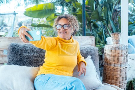 Photo for A woman uses her phone and relaxes alone in the garden, sitting on a sofa. Portrait of cheerful female writing on cellphone. Lady taking selfie. Mature young people relaxing - Royalty Free Image