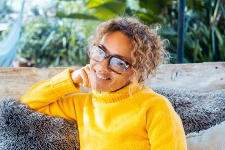 Happy blonde woman with glasses smiles sitting on sofa in garden near house