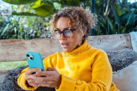 Photo for A woman uses her cell phone and relaxes alone in the garden, sitting on a sofa. Portrait of concentrated female writing on cell phone. Lady making a video call. Mature young people relaxing - Royalty Free Image