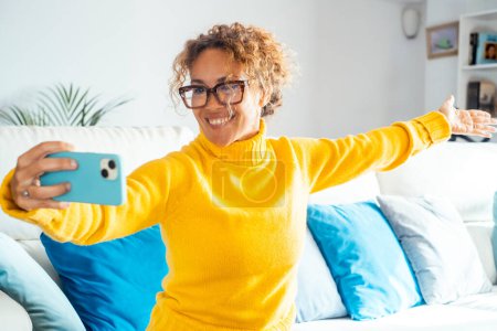 Foto de Mujer atractiva sentada en un sofá blanco en casa sonríe, toma selfie con su teléfono celular. Mujer feliz con suéter amarillo pasa un buen rato relajándose en casa. Concepto de ocio y relajación - Imagen libre de derechos