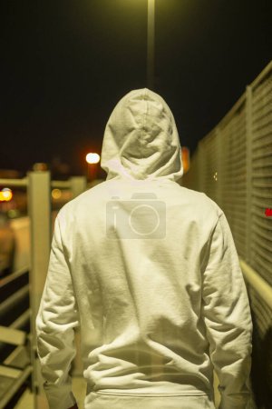 Photo for Back view of person with hoodie walking by night on the road with traffic cars on the street. Urban lifestyle people concept. Security and crime. Walk on a bridge - Royalty Free Image