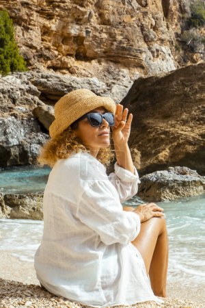 Téléchargez les photos : Touriste assise sur la plage tropicale profitant de vacances en plein air loisirs seul et portant un chapeau de paille. Les gens et les vacances d'été style de vie. Femme adulte se détendre dans la destination pittoresque - en image libre de droit