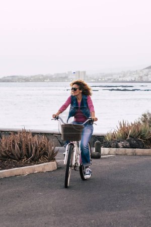 Foto de Señora montar en bicicleta solo en la calle con vista a la costa del océano. Actividad de ocio al aire libre mujer transporte verde. Personas y estilo de vida saludable. Concepto de turista de vacaciones - Imagen libre de derechos