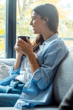 Young beautiful brunette woman with blissful facial expression alone on the couch with her bare feet on coffee table. Portrait of relaxed female resting at home.