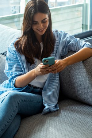 Happy girl checking social media while holding smartphone at home. Smiling young brunette woman using mobile phone app to play games, shop online, order delivery while relaxing on sofa. Scrolling.
