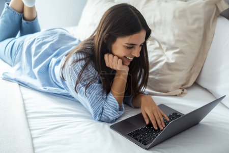  smiling woman lying down the bed in front of her laptop 