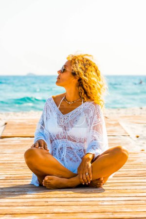 Photo for Beautiful woman with white swimsuit cover up on the tropical beach. Portrait of happy sitting blonde woman smiling at the beach. Curly tanned girl enjoying and relaxing at the seaside. - Royalty Free Image