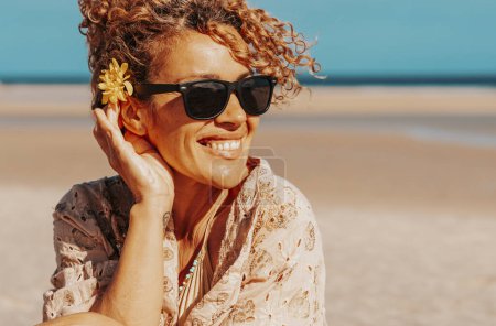 Photo for Front view of a beautiful woman sitting on the beach. Blonde woman with curly hair and yellow flower near ear relaxes looking and smiling. - Royalty Free Image