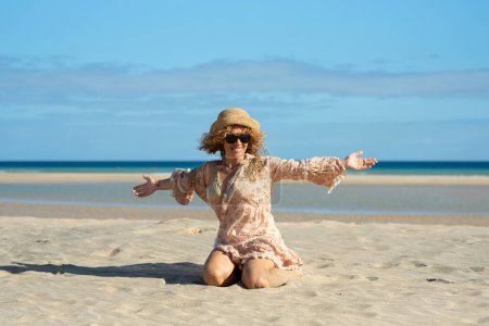 Foto de Vista frontal de una hermosa mujer con los brazos abiertos sentada en la playa. Mujer rubia con el pelo rizado soplando en el viento se relaja mirando y sonriendo. - Imagen libre de derechos