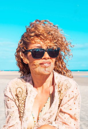 Téléchargez les photos : Front view of a beautiful woman sitting on the beach. Blonde woman with curly hair blowing in the wind relaxes looking and smiling. - en image libre de droit