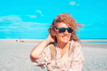Téléchargez les photos : Vue de face d'une belle femme assise sur la plage. Femme blonde aux cheveux bouclés soufflant dans le vent détend regarder et sourire. - en image libre de droit