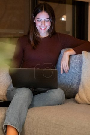 young woman using laptop by night at home sitting on the sofa searching on web and working late. Female people use wireless connection computer technology for work or leisure indoor activity