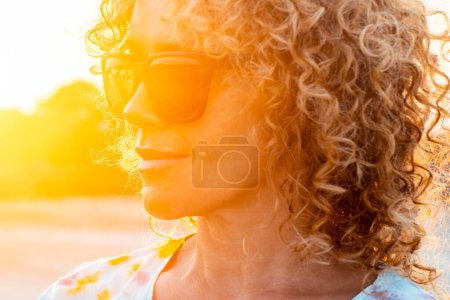 Photo for Outdoor portrait of curly woman smiling and wearing sunglasses with sunset backlight. Golden portrait of attractive lady in outdoors leisure activity alone. Tourist happy. Beautiful female - Royalty Free Image