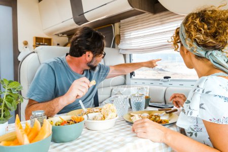 A happy couple spending time together having fun during vegetarian lunch inside a camper. People looking out the window pointing at the view with their finger thinking about the new destination.