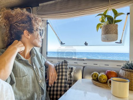 Téléchargez les photos : Femme bouclée avec des lunettes sourit en buvant du thé et en regardant par la fenêtre de son camping-car. Elle vacances à la maison sur roues et aventure printanière au bord de la mer. Mode de vie nomade concéder - en image libre de droit