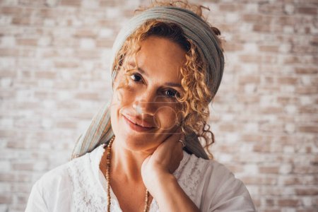 Happy curly woman smiles looking at the camera. Frontal portrait of a middle-aged lady with bricks indoor home background. One cute mid age female people smiling and looking at you. Cheerful lady
