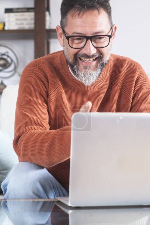 Photo for Bearded man with eyeglasses smiling and talking on video chat on laptop at home while sitting on sofa.Concept of people using mobile devices. - Royalty Free Image