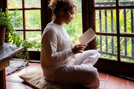 Photo for Serene woman reading a book at home sitting on the floor in total mindful chilling indoor leisure activity with nature green view outside the windows. People and study. Education. Relaxation - Royalty Free Image