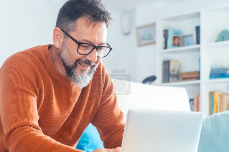  mature man alone at home using laptop sitting on sofa smiling and enjoying wireless technology and computer connection. Small business and alternative office, modern people lifestyle. Male beard