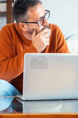  man sitting on sofa in front of an open laptop with thoughtful expression looking for a solution or idea. People and remote working lifestyle. Small business. Security password lost problems.